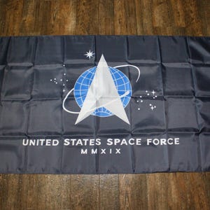 Op de afbeelding: Een zwarte vlag met het logo van de United States Space Force. Het logo is een witte ster in een blauwe cirkel met een witte baan eromheen. De tekst "UNITED STATES SPACE FORCE" en "MMXIX" is in wit onder het logo gedrukt.