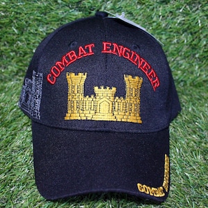 May include: A black baseball cap with the words "COMBAT ENGINEER" embroidered in red across the front. A gold castle emblem is centered below the text. The cap also features a gray castle design on the side and the word "COMBAT" embroidered on the brim.