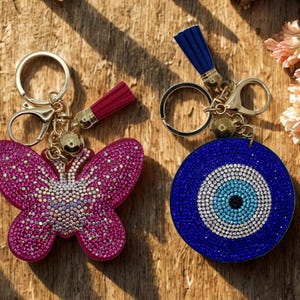 May include: Two keychains featuring gold-tone metal hardware, lobster clasps, and small tassels. One keychain is a butterfly shape covered in pink rhinestones, and the other is a circular evil eye design covered in blue and white rhinestones.