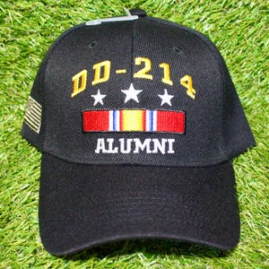 May include: Black baseball cap with the text "DD-214 ALUMNI" in yellow and white. The cap features a red, white, and blue ribbon design and a small American flag patch on the side. The cap is sitting on a green surface.