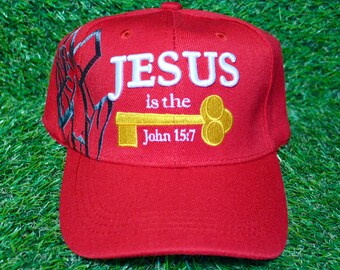 May include: A red baseball cap with the words "JESUS is the" and "John 15:7" embroidered in white and gold. The cap has a curved brim and a decorative design on the side.
