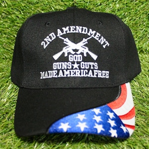 Peut inclure: Casquette de baseball noire avec texte brodé blanc : "2ND AMENDMENT GOD GUNS GUTS MADE AMERICA FREE." La visière présente un motif du drapeau américain. La casquette est posée sur un fond vert.