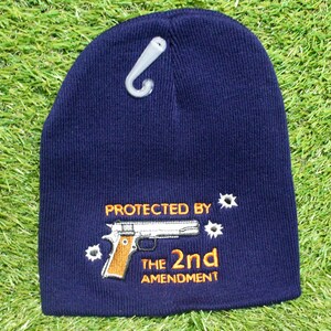 May include: Navy blue knit beanie with the embroidered text "PROTECTED BY THE 2ND AMENDMENT" and a graphic of a handgun. The beanie has a white plastic hang tag attached.