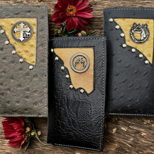 Puede incluir: Tres carteras de cuero en gris, negro y gris oscuro, cada una con una textura en relieve única. Cuentan con detalles metálicos decorativos, incluyendo cruces, caballos y revólveres, sobre un fondo de madera rústica.