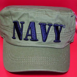 May include: Olive green military-style cap with a curved brim. The word "NAVY" is embroidered in dark blue across the front. The cap has a structured crown and a tag attached to the side.