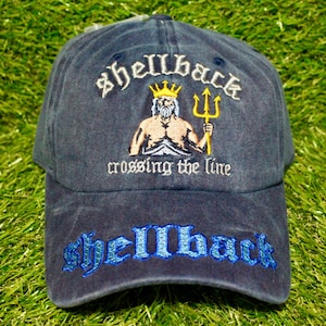 Peut inclure: Casquette de baseball bleu marine avec le mot "shellback" brodé en blanc et bleu. La casquette présente une image brodée d'une figure musclée avec une couronne et un trident, avec le texte "crossing the line".