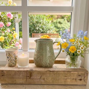Könnte beinhalten: Ein rustikales Holztablett hält eine grüne Keramikkrug mit floralem Relief, eine brennende Kerze, eine Blumentasse und ein Glas mit Blumen. Die Szene spielt sich vor einem Fenster mit Gartenblick ab.