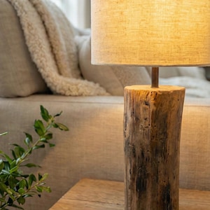 Puede incluir: Una l&aacute;mpara de mesa r&uacute;stica con una base cil&iacute;ndrica de tronco de &aacute;rbol. La l&aacute;mpara tiene una pantalla de lino beige y una varilla met&aacute;lica que conecta la pantalla a la base. La l&aacute;mpara est&aacute; sobre una mesa de madera, con un sof&aacute; y una planta al fondo.