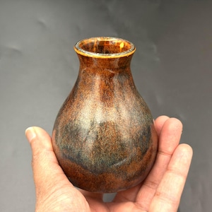 May include: A small, bulbous ceramic vase with a glossy finish. The surface features a mottled blend of deep brown, copper, and dark charcoal tones. The vase has a narrow neck and a slightly flared rim, showcasing a smooth, reflective glaze.