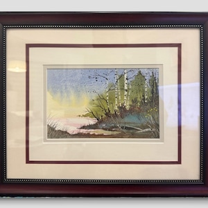 May include: Watercolour landscape painting of a tranquil lakeside scene, framed in a dark wood frame. The artwork depicts birch trees, green foliage, and a soft, colourful sky with pink and yellow hues. The painting is matted with a cream-coloured mat.
