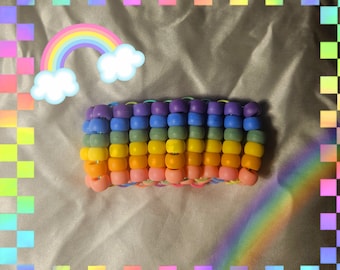 Könnte beinhalten: Ein farbenfrohes Perlenarmband in Regenbogenfarben, darunter Lila, Blau, Grün, Gelb, Orange und Rosa. Das Armband besteht aus kleinen, runden Perlen, die aneinandergereiht sind. Eine Regenbogen-Grafik befindet sich im Hintergrund.