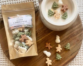 May include: A clear bag of "Golden Daze Cozy Christmas" wax melts with gingerbread men, snowmen, and Christmas trees. The melts are in a variety of colors, including white, tan, and green. The bag is on a wooden tray with a white ceramic bowl filled with similar melts.