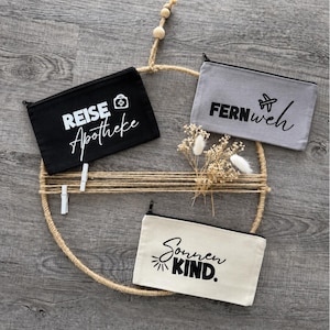 Könnte beinhalten: Drei rechteckige Reißverschlusstaschen aus schwarzem, grauem und cremefarbenem Canvas. Die schwarze Tasche trägt den weißen Schriftzug "REISE Apotheke" mit einem medizinischen Kreuz-Symbol. Die graue Tasche zeigt den schwarzen Text "FERNweh" mit einem Flugzeug-Symbol. Die cremefarbene Tasche zeigt den schwarzen Text "Sonnen KIND." mit einem Sonnensymbol.