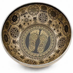 May include: A round metal singing bowl with a metallic gold finish and a black interior. The bowl features intricate black etched patterns including geometric shapes, lotus flowers, and a central pair of footprints surrounded by a decorative border.