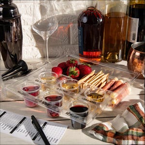 May include: A clear plastic tray with eight small cups for wine tasting. The cups are labeled with different wine varieties, including Malbec, Pinot Noir, Merlot, and Cabernet Sauvignon. There are also strawberries, crackers, and prosciutto on the tray.