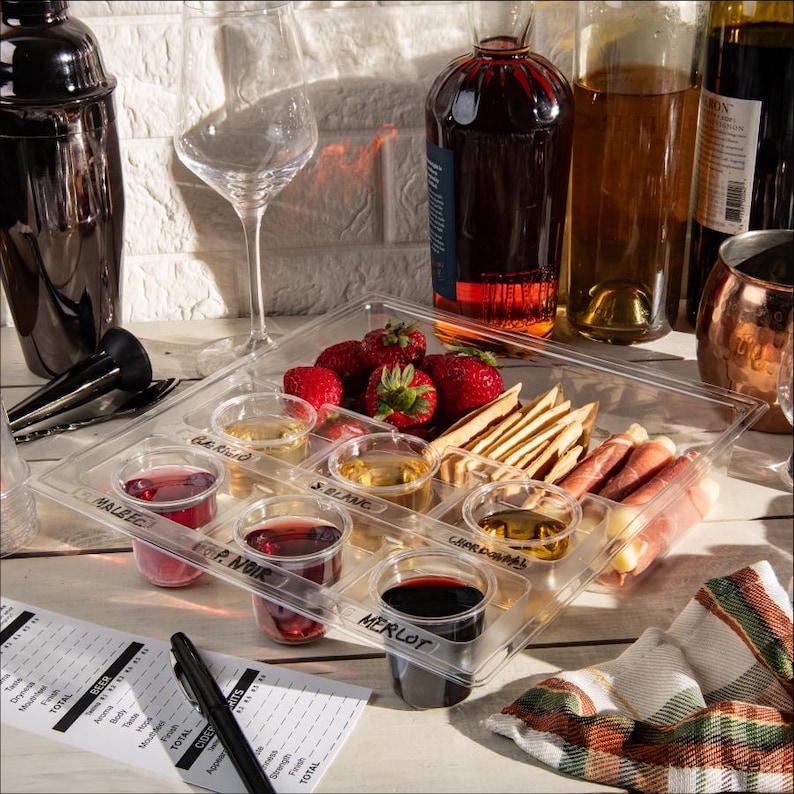 May include: A clear plastic tray with eight small cups for wine tasting. The cups are labeled with different wine varieties, including Malbec, Pinot Noir, Merlot, and Cabernet Sauvignon. There are also strawberries, crackers, and prosciutto on the tray.