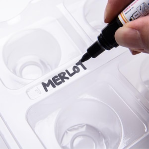 May include: A white plastic mold with six round cavities for making chocolate. The mold is labeled "MERLOT" in black marker.