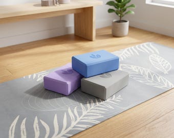 May include: Three yoga blocks in blue, purple, and gray, each with a leaf design, rest on a gray yoga mat with a white leaf pattern. A yoga strap and bench are in the background.