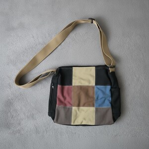 Op de afbeelding: Een patchwork schoudertas met een beige schouderriem. De tas heeft een zwarte basis met kleurrijke vierkante patches in rood, bruin, beige en blauw. Een klein vak met ritssluiting is aan de zijkant zichtbaar.