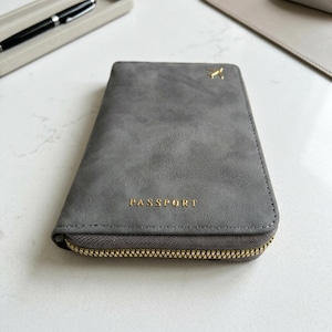May include: A gray passport holder with a gold zipper and the word "PASSPORT" in gold lettering. A small gold emblem is on the top right corner. The holder is made of a soft, suede-like material.