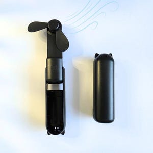 May include: A black handheld fan with a small, three-bladed fan on the top. The fan is attached to a long, cylindrical handle with a button on the side. The fan is next to a black, cylindrical case with a cat-shaped design on the top.
