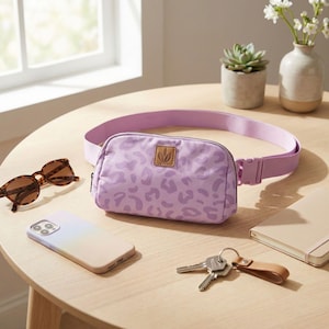 May include: A lilac fanny pack with a leopard print pattern, a matching adjustable strap, and a brown leather patch. The bag is on a wooden table with sunglasses, a smartphone, keys, and a notebook.