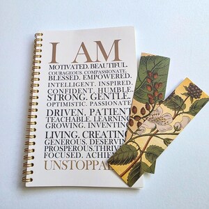May include: A white spiral bound journal with the words "I AM" in gold lettering. The journal is open to a page with a list of positive affirmations in gold lettering. A bookmark with a floral design is placed on the journal.