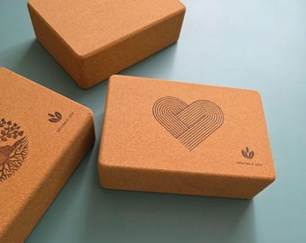 May include: Three rectangular yoga blocks made of light brown cork. One block features a heart design, another has a tree graphic, and the third is plain. The blocks are arranged on a teal surface, with the words "Neutral Zen" visible.