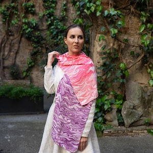 May include: A colourful scarf with a swirling pattern in pink and purple hues. The scarf is worn over a white dress. The background features a stone wall with green vines, providing a natural and stylish backdrop for the accessory.