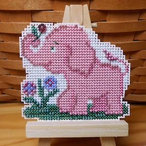 May include: A pink beaded cross-stitch of an elephant with a flower on its head standing in a field of green and blue flowers.