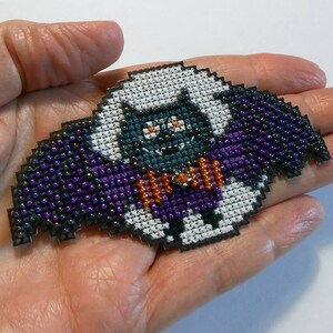 May include: A white and black cross-stitch bat with purple and orange wings. The bat has a black and white face with orange eyes and a black mouth.