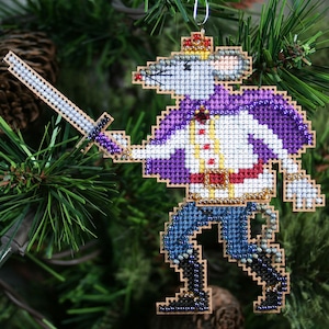 May include: A beaded Christmas ornament depicting a mouse dressed as a knight, holding a sword. The mouse is wearing a purple cape, a white shirt, a gold belt, and blue pants. The ornament is made of beads and is a festive decoration.