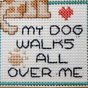 A close up of the square portion of the magnet. The phrase My Dog Walks All Over Me is back stitched with black floss, a heart is stitched with red floss above the word Dog and a paw print is stitched above the word over with light brown floss.