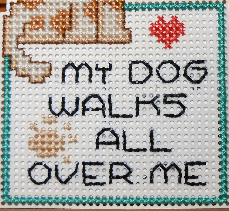 A close up of the square portion of the magnet. The phrase My Dog Walks All Over Me is back stitched with black floss, a heart is stitched with red floss above the word Dog and a paw print is stitched above the word over with light brown floss.