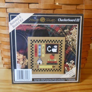 May include: A Mill Hill Checkerboard III cross stitch kit with a black and white checkered background. The kit includes a small schoolhouse, a cat, and books with the words "Red", "Blue", and "Yellow". The kit is 6.5 inches wide by 6 inches high.