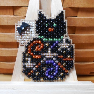 May include: A beaded Halloween decoration featuring a black cat with green eyes and a green collar. The cat is adorned with orange, purple, and silver beads. A small black bat is attached to the cat's side. The decoration is mounted on a wooden stand.