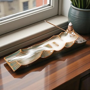 May include: A ceramic incense holder in the shape of a mountain range. The holder is primarily green with brown accents and a white smoke effect. An incense stick is lit, and a small incense cone sits on the peak.