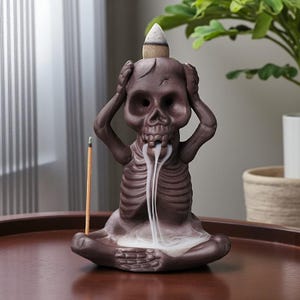 May include: A brown ceramic skull incense burner with a cone-shaped incense on top. The skull has its hands on its head and smoke is coming out of its mouth. An incense stick is placed in front of the skull.