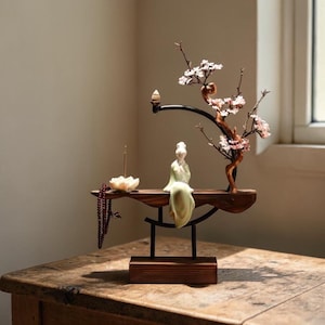 May include: A wooden zen garden sculpture featuring a seated figurine in a green robe, a flowering branch, and a lotus flower incense holder. The sculpture is on a wooden shelf with a dark metal support and a wooden base.
