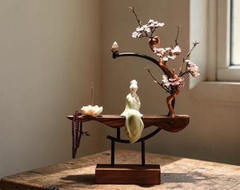 Op de afbeelding: Een houten zen-tuinbeeld met een zittend beeldje in een groene jurk, een bloeiende tak en een lotusbloem wierookhouder. Het beeld staat op een houten plank met een donkere metalen steun en een houten voet.