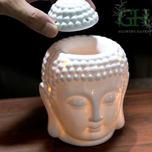 May include: White ceramic Buddha head oil warmer with a removable lid. The Buddha's face is serene, with closed eyes and a gentle smile. The warmer is lit from within, creating a warm glow. The product is from Glowing Haven.