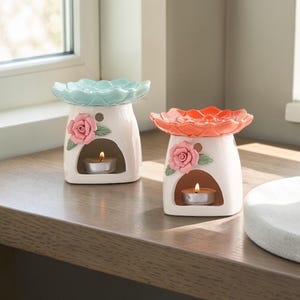 May include: Two ceramic oil warmers with floral designs. One has a light blue top and a pink rose, the other has an orange top and a pink rose. Each warmer has a small opening for a tea light candle.