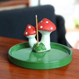 Può includere: Un porta incenso in ceramica con due figure di funghi rossi a pois bianchi, collegati alla base. I funghi poggiano su un piatto di ceramica verde, con un bastoncino di incenso acceso. Il supporto è progettato per l'aromaterapia.