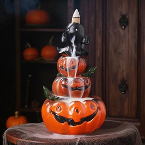 May include: A Halloween-themed incense burner featuring a stack of three orange jack-o'-lanterns with a black ghost figure on top. The burner emits a smoky effect, creating a spooky atmosphere. Green leaves are placed between the pumpkins.