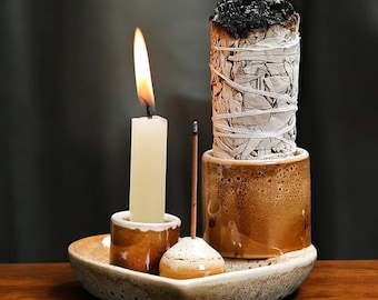 Può includere: Un portaincenso in ceramica con una candela accesa, un fascio di salvia che brucia e un bastoncino di incenso. Il supporto è marrone e beige, con una superficie strutturata. La fiamma della candela è gialla e arancione.