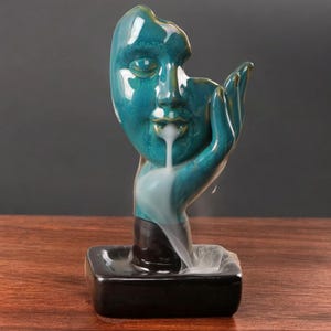 May include: A teal blue ceramic incense burner shaped like a face with a hand holding the face. The incense smoke flows out of the mouth and down the hand.