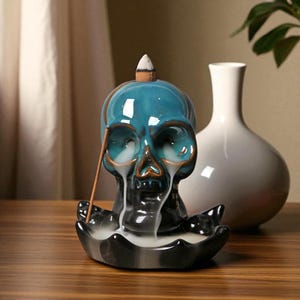 May include: A teal ceramic skull incense burner with a brown accent. The skull-shaped burner has smoke emanating from the eye sockets and mouth. A white incense cone sits on top. A white vase is in the background.