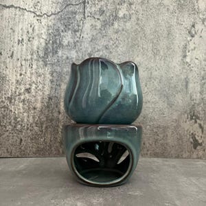Puede incluir: Un quemador de aceite de cerámica azul verdoso con forma de tulipán y una base decorativa con calados.