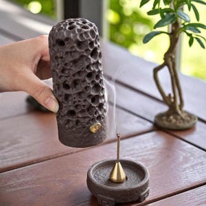 May include: A dark brown incense holder with a porous design is held above a matching incense burner. A gold-colored incense stick is lit, with smoke rising. A small bonsai tree is in the background.