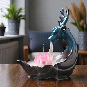 May include: A teal and grey ceramic incense burner shaped like a deer. Smoke flows from the deer's mouth, above a pink lotus flower. An incense stick is inserted into the flower. The burner sits on a wooden table, with a blue cushion in the background.